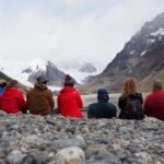 Seitsemän selin istuvaa ihmistä kivikkoisella rannalla katselemassa vuoria, valokuva.